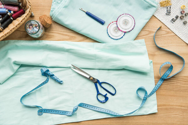 верхний вид рабочего места швеи на стол с тканью, ножницами, иглами, измерительной лентой и кнопками
