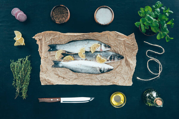 top view of gourmet fish with lemon slices on baking paper and spices on black