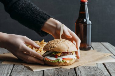 cropped view of female hands with hamburger, french fries and bottle of beer on baking paper on wooden table clipart