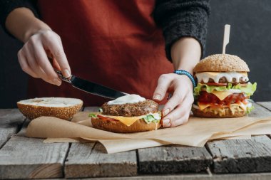 cropped view of girl in apron making tasty homemade cheeseburgers on baking paper on wooden tabletop clipart