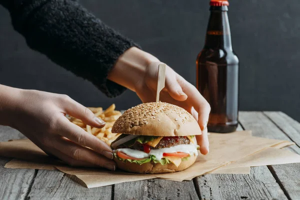 木製のテーブルに紙を焼くのハンバーガー フライド ポテト ビールの瓶を持つ女性の手のビューをトリミング — ストック写真