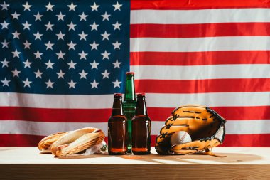 close-up view of beer bottles, hot dogs, leather glove and baseball ball on wooden table with us flag behind clipart