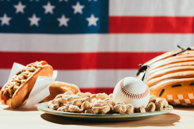 close-up view of baseball ball on plate with peanuts, leather glove and hot dogs on table with us flag behind clipart