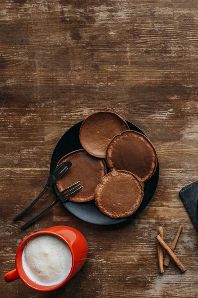 木桌上有煎饼和卡布奇诺杯的黑盘子的顶端视图 — 图库照片