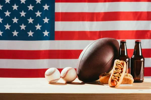 close-up view of hot dogs, beer bottles and balls on wooden table with us flag behind 