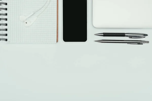 top view of notebook with earphones, smartphone, laptop and pens on white