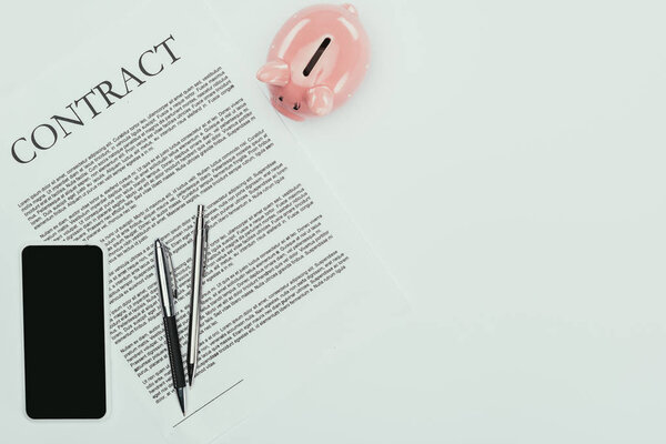 top view of smartphone with blank screen, contract with pens and piggy bank on white 
