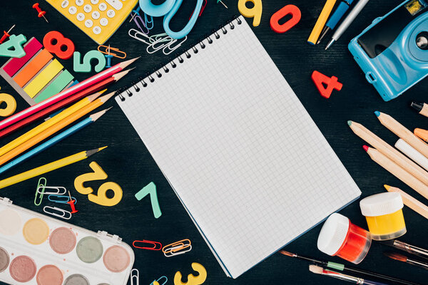 Top view of composition of colorful school supplies with blank notebook isolated on dark board background