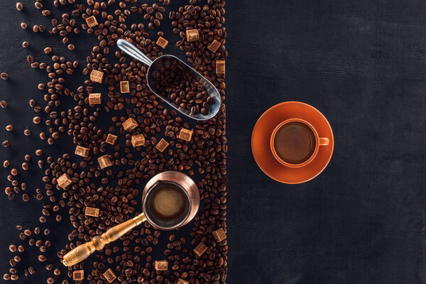 top view of cup of coffee with saucer, roasted coffee beans, brown sugar, coffee pot and scoop on black