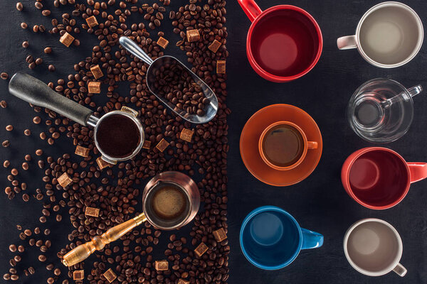 top view of roasted coffee beans, scoop, coffee pot, coffee tamper and cup of coffee with empty cups on black