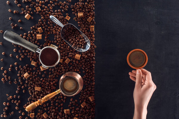 partial view of person holding cup of coffee and roasted coffee beans with coffee pot, scoop and sugar 