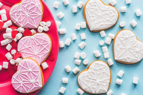 top view of glazed heart shaped cookies on pink plate with white marshmallow isolated on blue