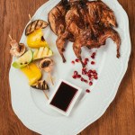Grilled chicken laying on plate with fried vegetables on skewer near saucer over wooden surface