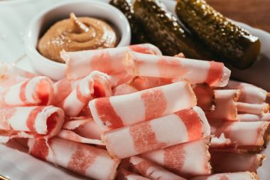 Close-up view of sliced bacon served with sauce and pickled cucumbers on plate