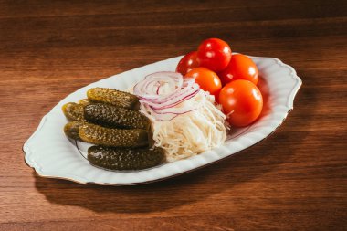 marinated salt cucmbers, sour cabbage with onion rings and raw tomatos laying on plate over wooden surface  clipart
