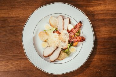 Ahşap masa üzerinde beyaz tabakta üstten görünüm Sezar salatası tavuk eti, domuz eti, yumurta ve peynir sosu ile servis