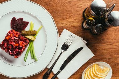 Top view of Vinegret salad served with green onion and pickled cucumber on white plate with condiment dispensers on wooden table clipart