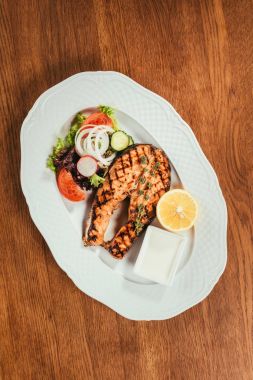 Top view of grilled salmon fish pieces with lemon, herbs and salad on white plate on wooden table clipart