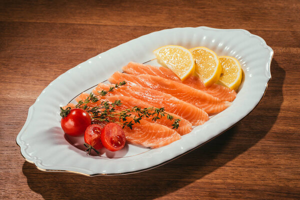 Salmon fish pieces with lemon, herbs and tomatoes on white plate on wooden table