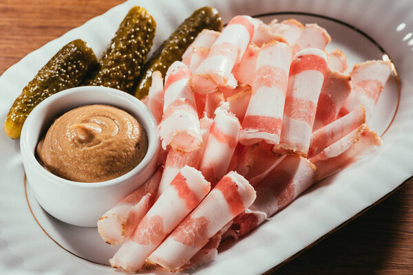 close up of cold-boiled pork with mustard in saucer on white plate over wooden surface  