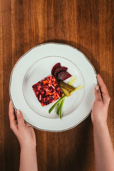 Top view of female hands holding plate with Vinegret salad served with green onion and pickled cucumber on wooden table
