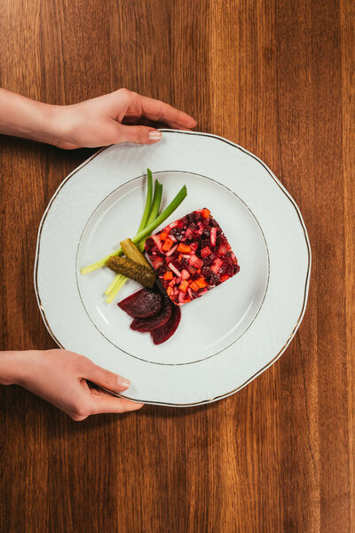 Top view of female hands holding plate with Vinegret salad served with green onion and pickled cucumber on wooden table