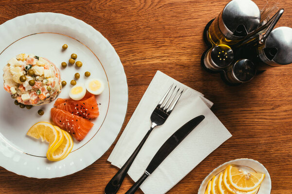 Русский салат на тарелке с разбросанным горохом, вареными яйцами и ломтиками рыбы на столе с вилкой и ножом на салфетке
