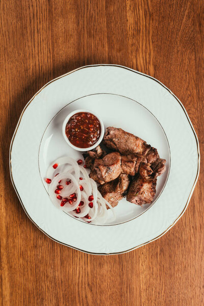 Top view of grilled meat with onion, sauce and pomegranate seeds on white plate on wooden table