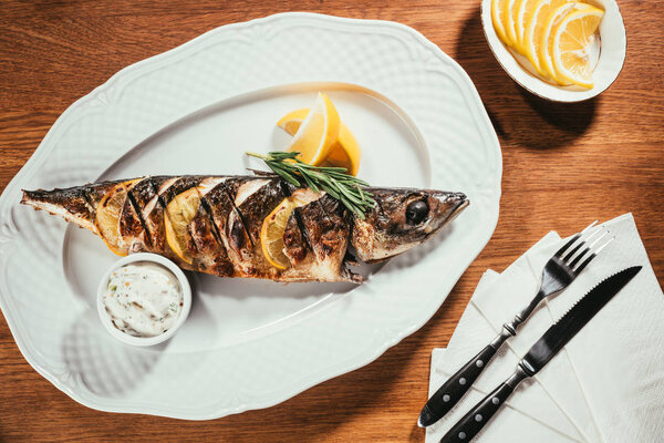 Baked fish with lemon and herbs on white plate with sauce on wooden table