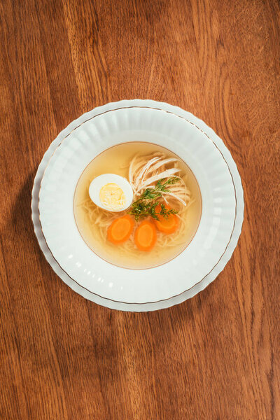 Top view of chicken soup with vegetables and egg in white plate on wooden table