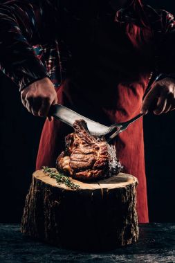 cropped shot of chef in apron holding fork and knife while slicing delicious grilled ribs on wooden stump clipart