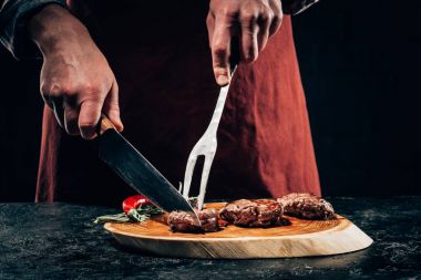 cropped shot of chef in apron with meat fork and knife slicing gourmet grilled steaks with rosemary and chili pepper on wooden board  clipart