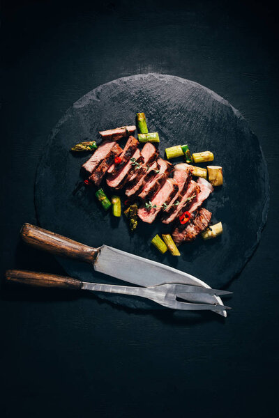 top view of delicious sliced grilled meat with asparagus, meat fork and knife on black slate board 