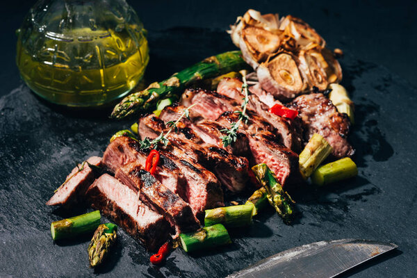 close-up view of tasty sliced grilled meat with asparagus, oil and spices on black
