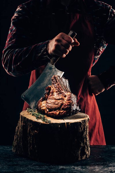 cropped shot of chef in apron holding meat knife and delicious grilled ribs on wooden stump 