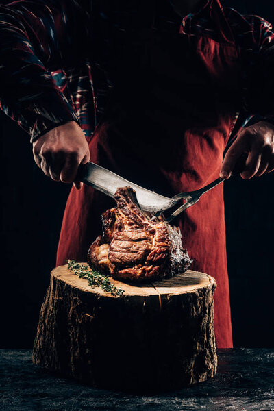 cropped shot of chef in apron holding fork and knife while slicing delicious grilled ribs on wooden stump