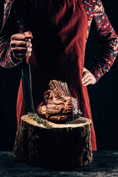 cropped shot of chef in apron standing with meat knife and delicious grilled ribs on wooden stump 