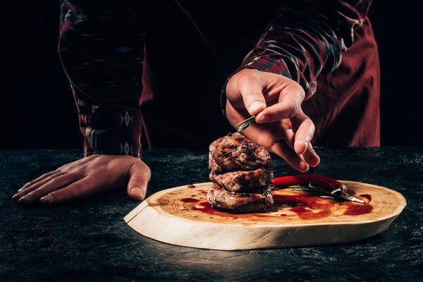 close-up partial view of chef putting rosemary on grilled steaks with chili pepper and sauce on wooden board