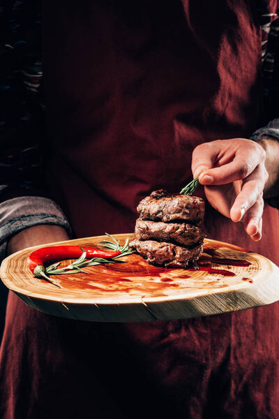 mid section of chef in apron holding wooden board with delicious grilled meat, chili pepper, sauce and rosemary 