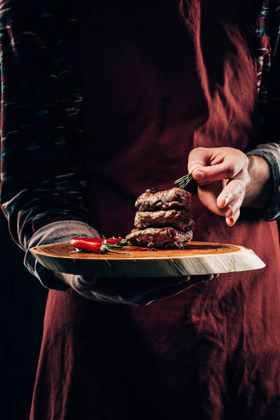 обрезанный выстрел шеф-повара в фартуке держа деревянную доску с вкусным мясом гриль, чили перец и розмарин
 