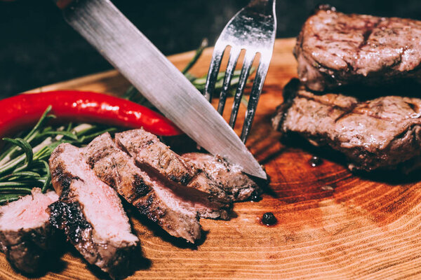 крупный план нарезанного мяса на гриле с розмарином и перцем чили с вилкой и ножом на деревянной доске
