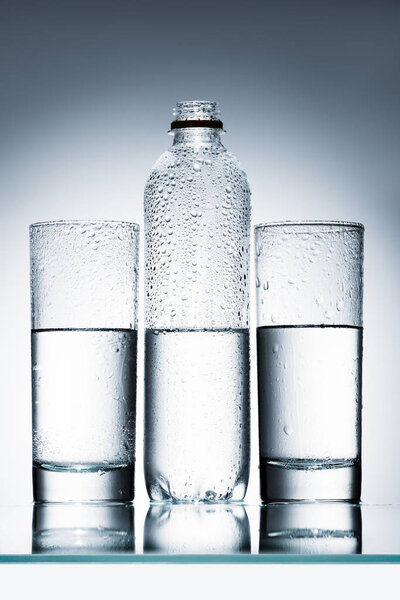 пластиковая бутылка и стаканы чистой воды в ряд на отражающей поверхности
