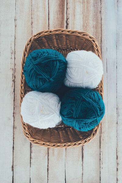 top view of white and blue knitting wool balls in wicker basket on wooden background