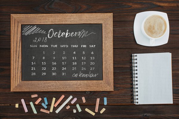 top view of arranged cup of coffee, empty notebook, colorful chalks and chalkboard with october calendar