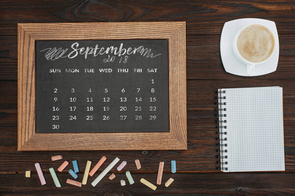 top view of arranged cup of coffee, empty notebook, colorful chalks and chalkboard with september calendar