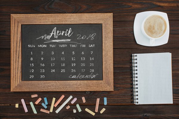top view of arranged cup of coffee, empty notebook, colorful chalks and chalkboard with april calendar