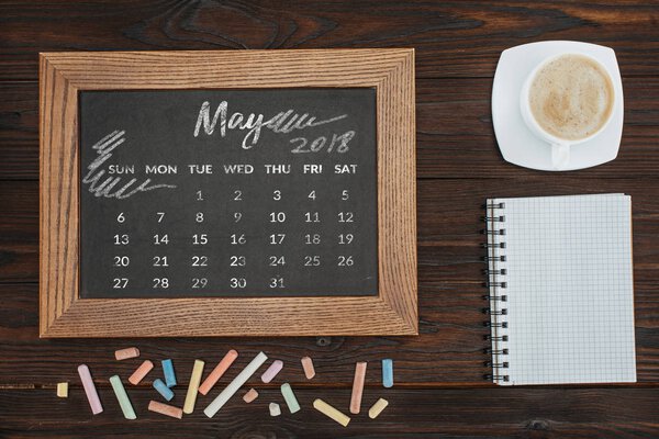 top view of arranged cup of coffee, empty notebook, colorful chalks and chalkboard with may calendar
