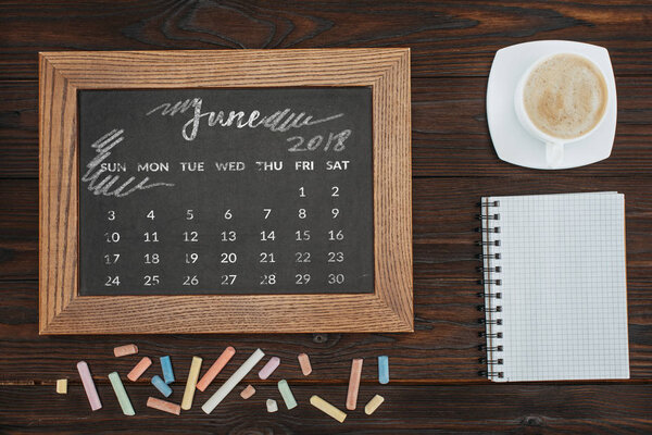 top view of arranged cup of coffee, empty notebook, colorful chalks and chalkboard with june calendar