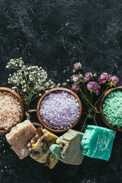 top view of natural soap, dried flowers and sea salt in wooden bowls on black marble surface, spa concept