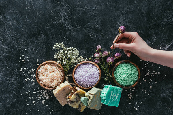 cropped view of female hand with natural homemade soap, dried flowers and sea salt for spa on black marble surface